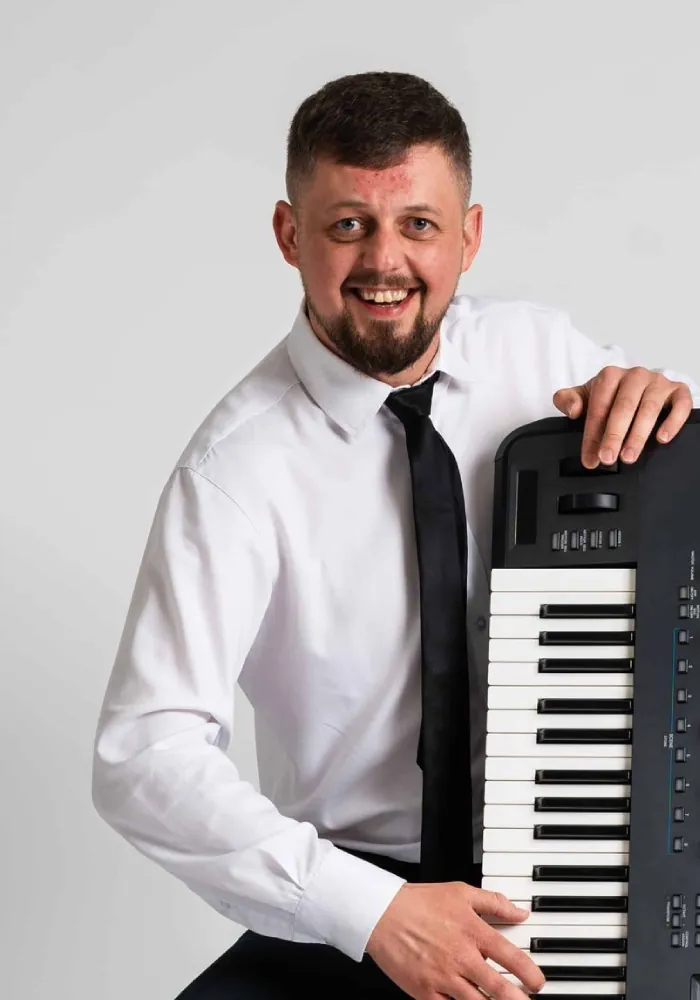 Szeroko uśmiechnięty mężczyzna w białej koszuli i czarnym krawacie, trzymający pionowo instrument klawiszowy.