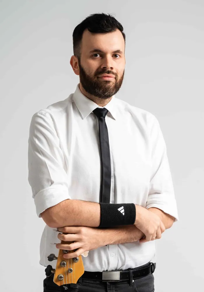 Brodaty mężczyzna ze skrzyżowanymi ramionami w białej koszuli i czarnym krawacie, trzymający gryf gitary.