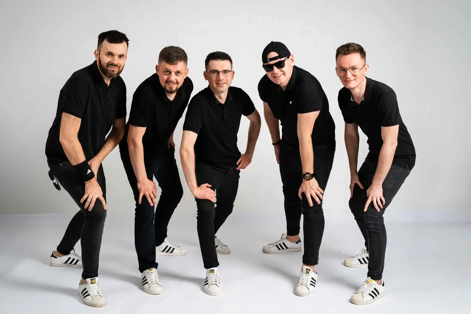 Zgrana ekipa pięciu muzyków w czarnych koszulkach polo i białych trampkach, pozująca w dynamicznej grupie.