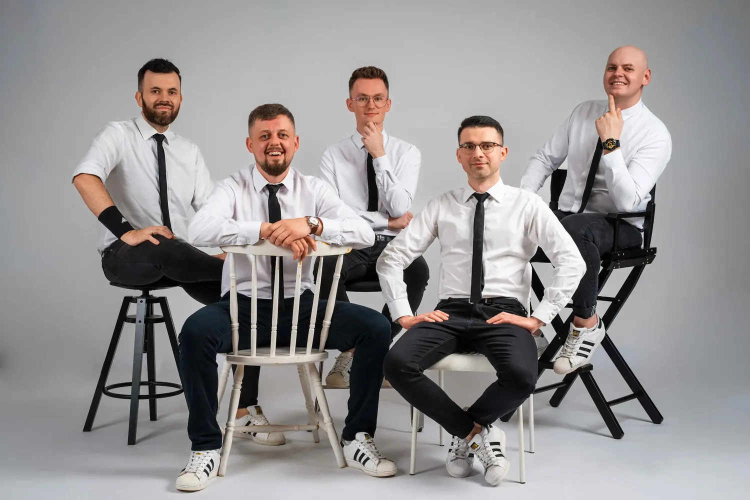 Pięciu uśmiechniętych członków zespołu w eleganckich białych koszulach i krawatach, pozujących wspólnie na krzesłach.